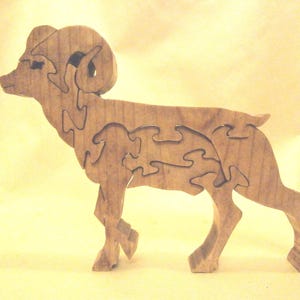 May include: A light brown wooden puzzle shaped like a bighorn sheep. The figure is composed of several interlocking pieces that fit together to form the complete animal silhouette, showing the head, curved horns, body, and four legs.