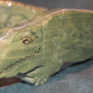 May include: A green wooden jigsaw puzzle shaped like an alligator. The puzzle pieces are cut into irregular shapes, and the alligator features a dark brown wood-burned outline for the eye and mouth. The surface has a visible wood grain texture.