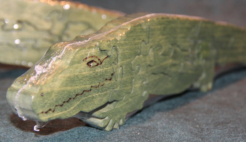 May include: A green wooden jigsaw puzzle shaped like an alligator. The puzzle pieces are cut into irregular shapes, and the alligator features a dark brown wood-burned outline for the eye and mouth. The surface has a visible wood grain texture.