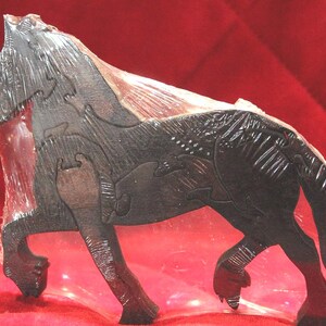 May include: A dark brown wooden horse puzzle featuring interlocking pieces with carved details on the mane, tail, and legs. The horse is depicted in a standing position with a textured surface finish, encased in a clear protective plastic film.