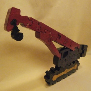 May include: A wooden puzzle of a red and black construction crane. The crane has a long arm and a hook on the end. The crane is on a black base with gold tracks.