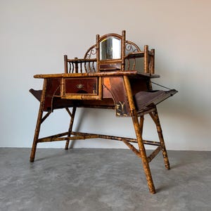 May include: Antique bamboo vanity desk with a mirror, drawers, and side shelves. The desk has a dark brown finish with gold accents and a unique, open-legged design. The mirror is framed with decorative metalwork.