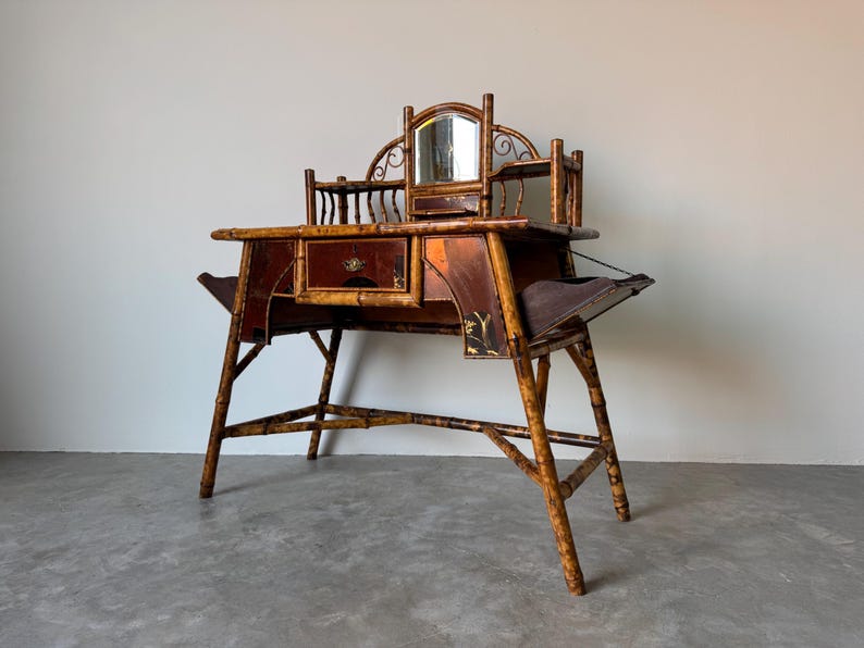 May include: Antique bamboo vanity desk with a mirror, drawers, and side shelves. The desk has a dark brown finish with gold accents and a unique, open-legged design. The mirror is framed with decorative metalwork.