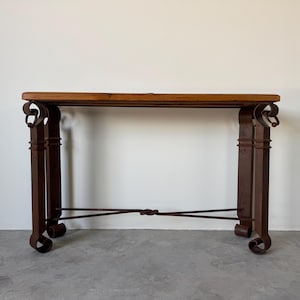 May include: A rustic console table with a rectangular wooden top and ornate wrought iron legs. The legs are dark brown with decorative scrollwork and a connecting bar. The table is approximately 48 inches long.