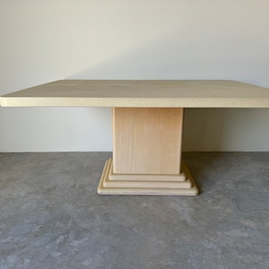 May include: A square, off-white dining table with a pedestal base. The base has a layered, stepped design.