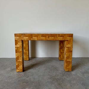 May include: A square, wooden end table with a woven pattern. The table has a light brown finish and is made of natural materials.