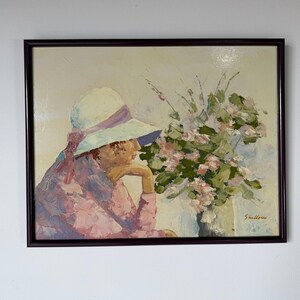 May include: An oil painting in a dark brown frame depicting a person wearing a white hat with a purple ribbon and a pink textured shirt. The person rests their chin on a hand next to a large bouquet of pink flowers and green leaves. Signed Shallow.