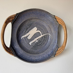 May include: A round, blue ceramic serving platter with woven handles. The platter has a white abstract design in the center. The handles are made of woven, light brown material. The platter is a decorative piece.