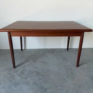 Puede incluir: Una mesa de comedor rectangular de madera con un acabado marrón cálido y veta de madera visible. La mesa presenta una superficie lisa y plana con bordes redondeados y cuatro patas cilíndricas cónicas que se extienden desde las esquinas.