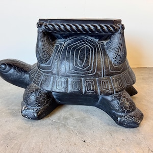 May include: Black turtle-shaped garden stool. The stool has a flat top with a rope-like border and is supported by a turtle-shaped base. The turtle's shell features detailed carved patterns. The stool is made of a dark material.