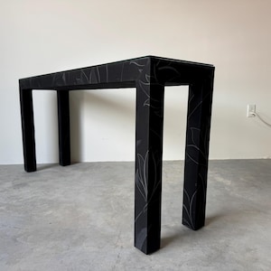 May include: A black console table with a glass top and four square legs. The table's surface and legs are decorated with a subtle, light-colored leaf pattern. The table is approximately 30 inches tall.