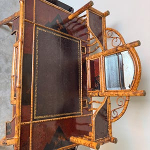 May include: An antique bamboo and leather desk with a mirror. The desk features a dark brown leather writing surface with gold trim, and a decorative mountain and floral design. The frame is made of bamboo with a natural finish.
