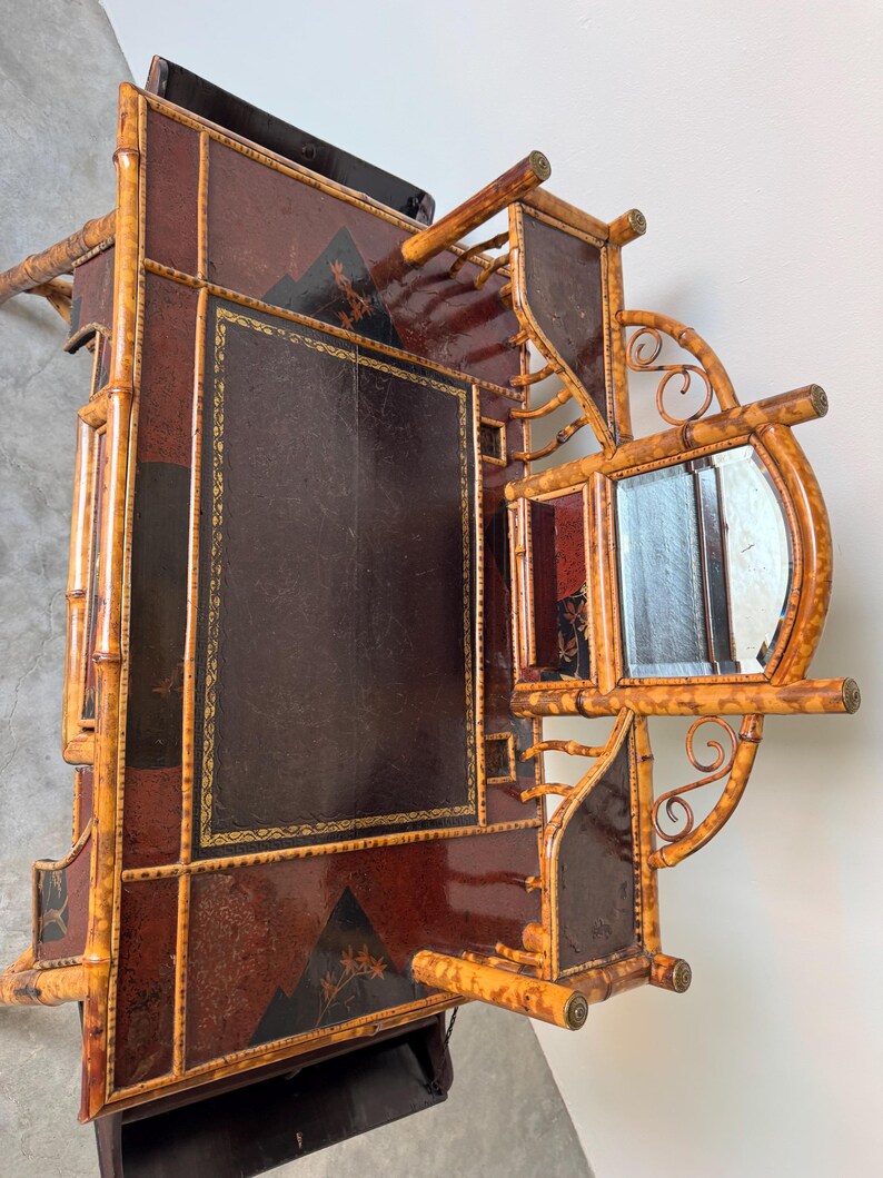 May include: An antique bamboo and leather desk with a mirror. The desk features a dark brown leather writing surface with gold trim, and a decorative mountain and floral design. The frame is made of bamboo with a natural finish.