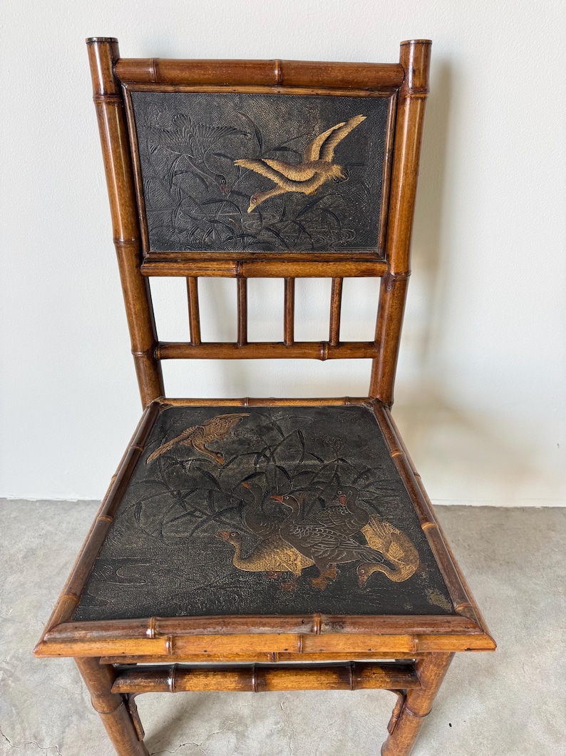 May include: A vintage bamboo chair with a dark seat and back featuring embossed bird designs in gold and brown. The chair has a natural wood finish and a classic, elegant design, suitable for home decor.