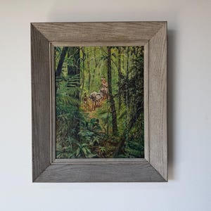 May include: Framed print depicting a lush jungle scene with figures and animals on a path. The artwork features vibrant greens and browns, with a light-colored path winding through the dense foliage. The frame is a light gray color.