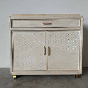 May include: White wooden cabinet with a woven pattern and gold hardware. The cabinet has a single drawer and two doors. The cabinet is on wheels.