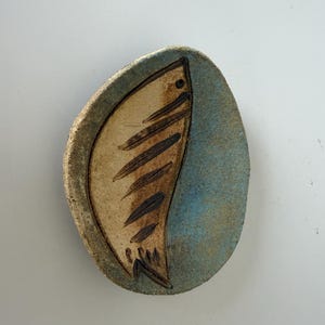 May include: An oval-shaped ceramic dish with a textured, sandy beige rim. The center features a stylized fish illustration in dark brown on a smooth, light beige background, contrasted by a textured, mottled blue glaze on the right side.
