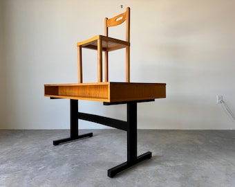 May include: A unique, mid-century modern desk with a built-in shelf and a small wooden chair on top. The desk has a black base and a light brown wooden top. The chair is also light brown.