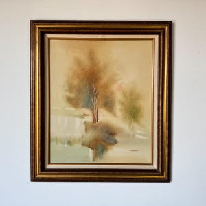 May include: A framed painting of a landscape with a single tree in the center. The painting is done in muted tones of brown, green, and white, and the background is a soft blur.