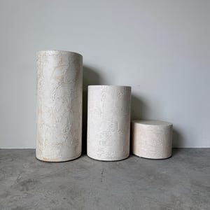 May include: Three cylindrical pedestals in varying heights. The pedestals have a textured, off-white finish with subtle beige accents. They are arranged on a gray concrete floor against a plain white wall, suitable for displaying art or decor.