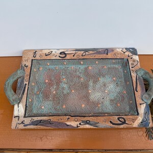 May include: Rectangular ceramic tray with a turquoise and copper interior, adorned with a wavy pattern and small dots. The tray has a brown border with black abstract designs and turquoise handles. A decorative piece.