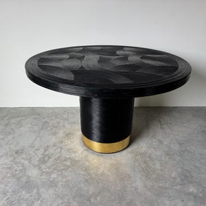 May include: A round black table with a textured top and a cylindrical base. The base has a gold-colored band at the bottom. The table is made of woven material and is suitable for a living room or dining area.