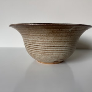 May include: A beige ceramic bowl with a wide, flared rim and a textured, horizontal striped pattern. The bowl has a dark brown rim and a light brown base. The bowl is likely handmade and suitable for serving food.