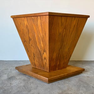 May include: A wooden table with a square top and a triangular base. The table is made of light brown wood with a natural finish.