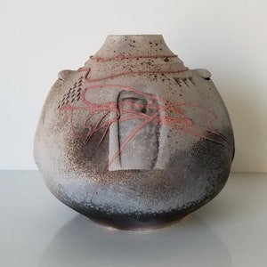 May include: A large, round, gray ceramic vase with a textured surface and red accents. The vase has a wide mouth and a narrow base.