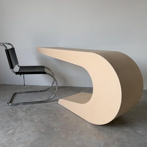 May include: A beige desk with a continuous, curved C-shaped silhouette and a textured finish, paired with a chair featuring a black seat and backrest supported by a polished chrome tubular metal frame that forms a cantilevered base structure.