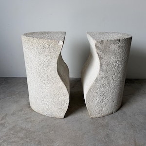 May include: A pair of off-white, textured stone pedestals featuring a curved, concave side profile and a flat, triangular top surface. The exterior surfaces exhibit a rough, porous finish with vertical striations throughout the structure.