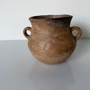 May include: A small, rustic, brown ceramic pot with two loop handles. The rim is dark brown, and the body has a textured, aged appearance. The pot sits on a white surface, suggesting a decorative or functional item.