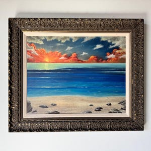 May include: A framed painting of a seascape with a vibrant orange and red sunset over a blue ocean. The painting depicts a sandy beach with rocks in the foreground.