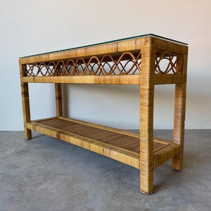 May include: A natural rattan console table with a glass top. The table features a lower shelf and decorative woven detailing on the front. The frame is made of light brown rattan, and the table is suitable for a living room or entryway.