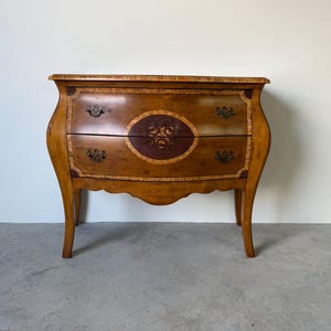 May include: A two-drawer wooden chest with a curved front and legs. The chest is stained a warm brown with decorative floral accents and ornate hardware. An oval-shaped floral design is centered on the front. The chest is approximately 30 inches wide.