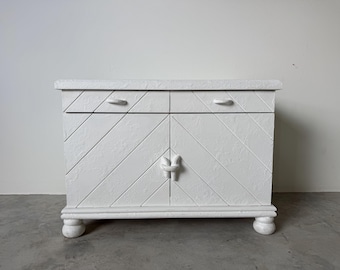 May include: White painted cabinet with two drawers and two doors. The cabinet has a textured surface with diagonal lines and a knot-shaped handle. It stands on four rounded feet.