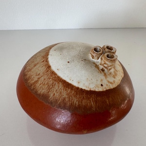 May include: A handcrafted ceramic vessel with a rounded shape. The base is a deep reddish-brown, transitioning to a lighter brown band, and topped with a speckled white area. Three small, cylindrical protrusions are on top.
