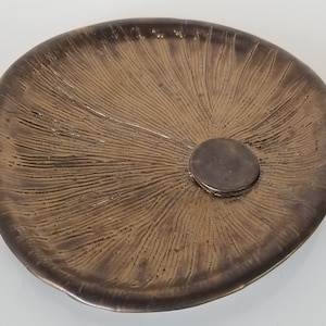 May include: A brown ceramic dish with a textured surface and a circular indentation in the center. The dish has a slightly irregular shape.