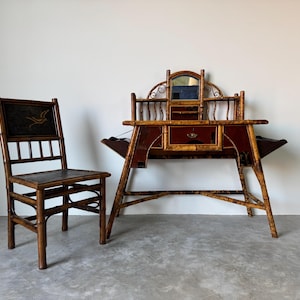 May include: Antique bamboo desk and chair set. The desk features a red and gold finish with a mirror and drawers. The chair has a dark seat and back with a decorative design. A unique vintage furniture set.