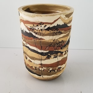 May include: A handcrafted ceramic vase with a cylindrical shape. The vase features horizontal bands of varying earth-tone colors, including beige, brown, and black. Delicate, stylized plant silhouettes are painted on the front. A decorative piece for home decor.
