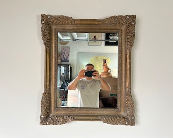 May include: A large, ornate, gold-toned mirror with a heavily carved frame. The mirror is reflecting a person taking a selfie.