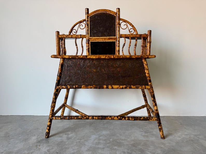 May include: An antique bamboo desk with a dark brown, textured surface. The desk has a high back with a decorative arch and a small mirror. The frame is made of bamboo with a natural finish.