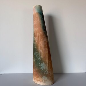 May include: A tall, slender, conical ceramic vase featuring a mottled glaze with blended tones of earthy brown, terracotta, and muted teal. The surface has a textured, weathered appearance with a smooth, rounded opening at the top.