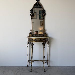 May include: A tall, narrow console table with an integrated arched mirror. The frame is made of dark metal with gold-toned accents, featuring a semi-circular marble top. The mirror has a pointed top and decorative metal scrollwork details.