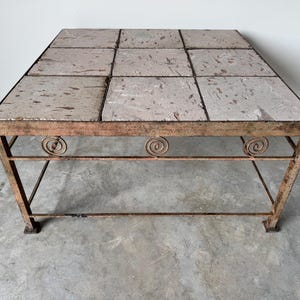 May include: A square coffee table with a tiled top made of light brown and pink stone. The table has a metal frame with a simple design and a lower shelf.