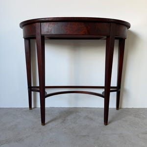 May include: A semi-circular console table crafted from dark mahogany wood with a polished finish. The table features four tapered legs connected by a curved lower stretcher, with small metallic caps at the base of each leg and a smooth top surface.
