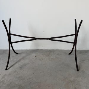 May include: A dark brown metal table base with a unique design. The base features curved legs and a cross-braced support structure. The metal has a textured, rough appearance, and the overall design is modern and industrial.