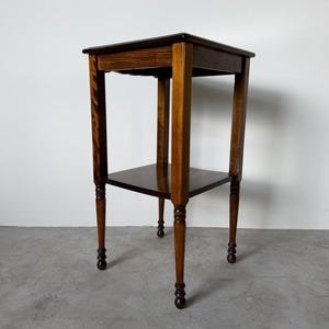 May include: A square wooden side table with a dark brown finish, featuring a flat top surface and a lower shelf. The table stands on four slender legs that have decorative turned details near the base, providing a stable and elevated structure.