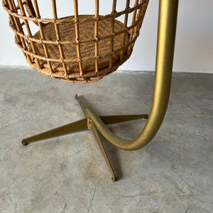 May include: A wicker hanging chair with a gold metal stand. The chair is suspended from the stand by a curved metal rod.