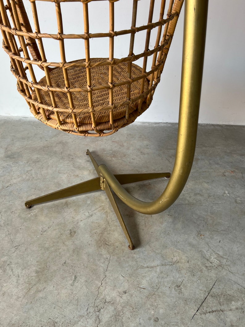 May include: A wicker hanging chair with a gold metal stand. The chair is suspended from the stand by a curved metal rod.